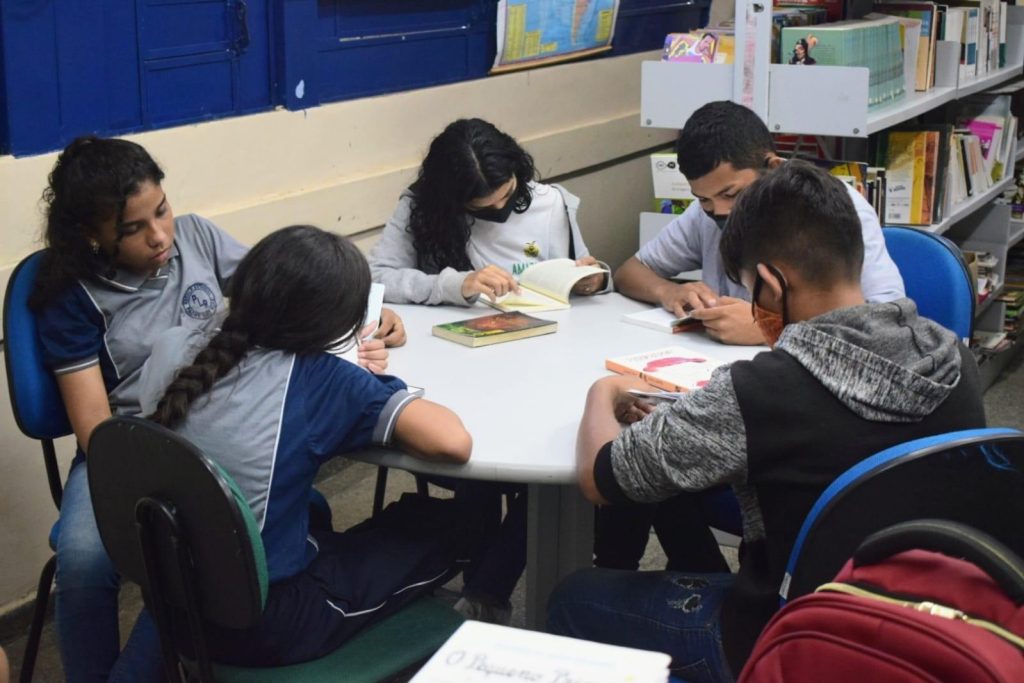 Alunos sentados em uma mesa redonda, localizada em uma biblioteca, lendo exemplares de livros