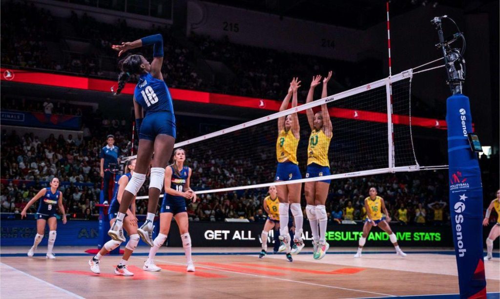 Jogadoras de vôlei disputando bola na rede. De um lado, jogadora de azul, da Itália, do outro do Brasil, de verde e amarelo e shorts azuis