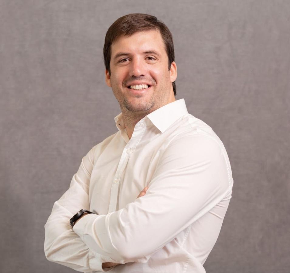 Homem branco usando uma camisa social branca cruza os braços e sorri