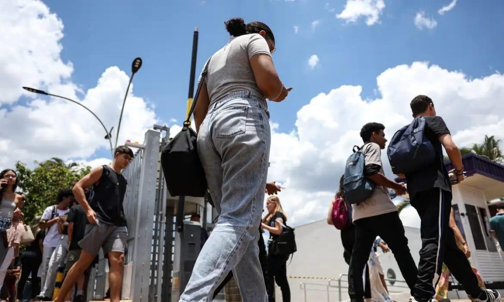 Imagem de uma mulher de costas. Ela veste uma calça jeans e uma blusa cinza. Tem os cabelos presos e olha o celular. Ao lado, uma mochila preta. Ao fundo, outras pessoas andam para entrar em algum lugar