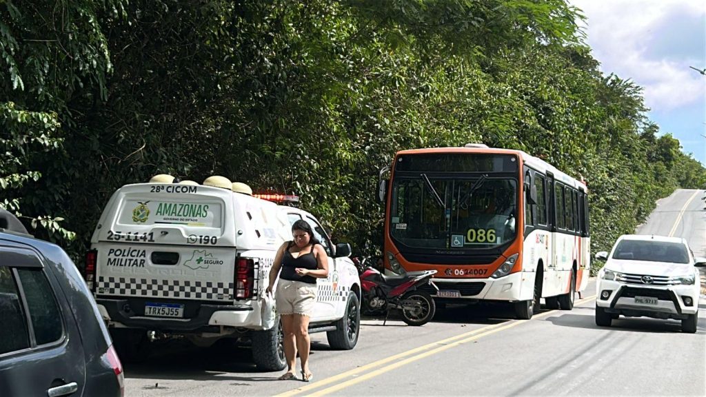 Imagem de um ônibus vermelho e branco onde tem a placa 086. Ele está parado no meio-fio. À frente, está um carro da Polícia Militar do Amazonas. Uma pickup passa ao lado do ônibus na rua