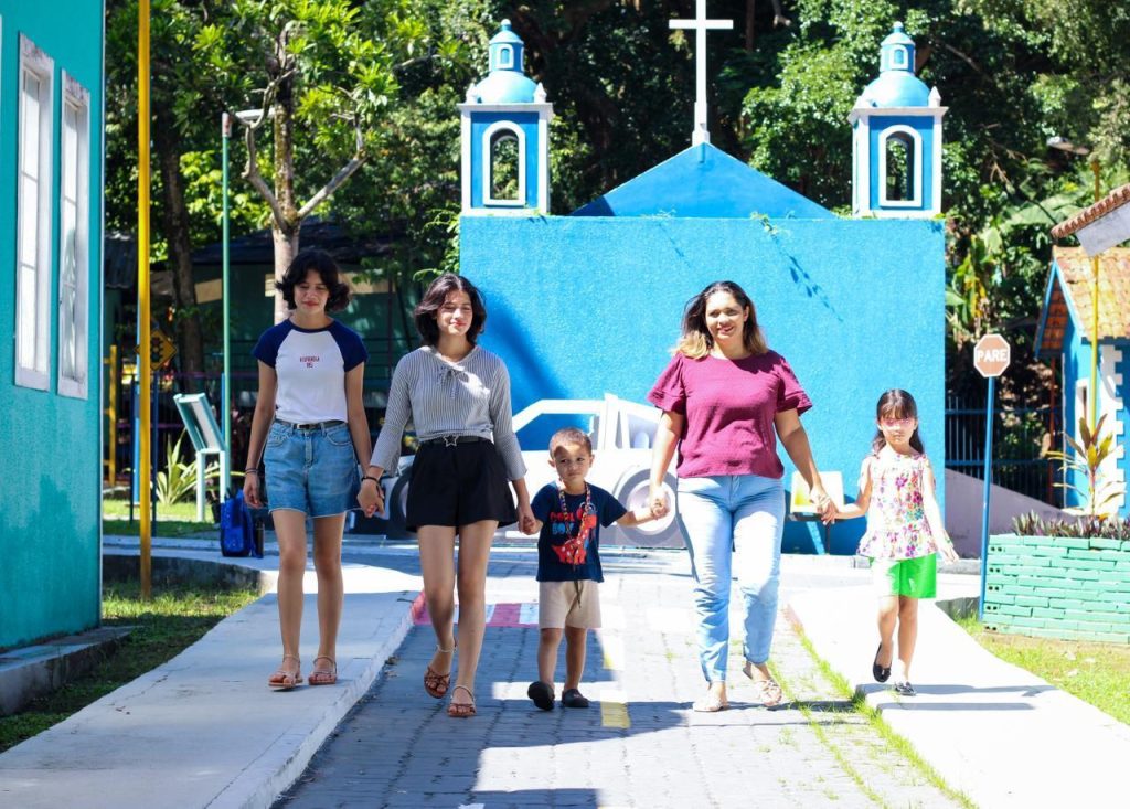 Imagem de uma mulher segurando a mãos dos filhos. A mulher está usando uma camisa rosa e jeans é baixa e tem cabelos tingidos de loiro. À esquerda dela está uma menina de vestido branco florido. À direita, um menino de camisa preta e bermuda de cor clara. Segurando a mão dele, duas adolescentes gêmeas de cabelos curtos. Uma usa uma camisa branca e preta e saia jeans, a outra, um camisa cinza e  saia preta