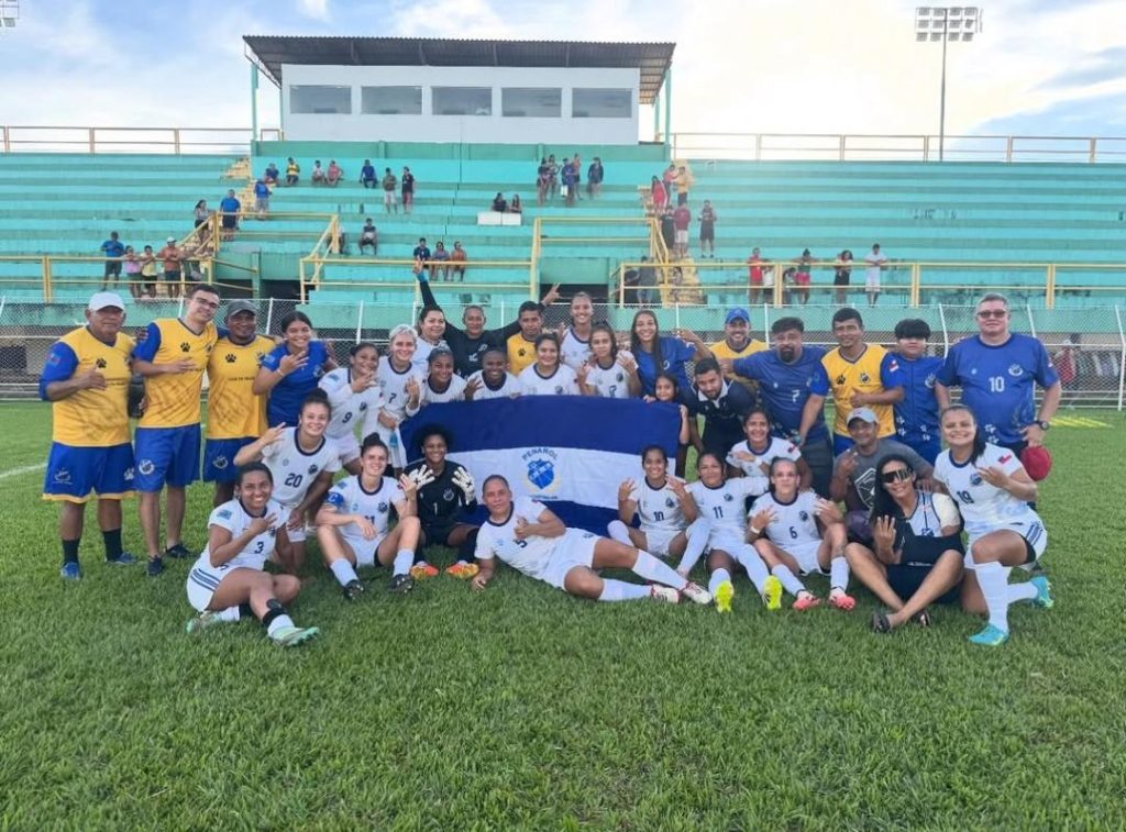 Equipe do Penarol Feminino. Jogadoras estão sentadas no gramado  rodeando uma bandeira do clube.  Elas usam uniforme branco, com detalhes em azul. A comissão técnica usa uniformes azuis com detalhes amarelos