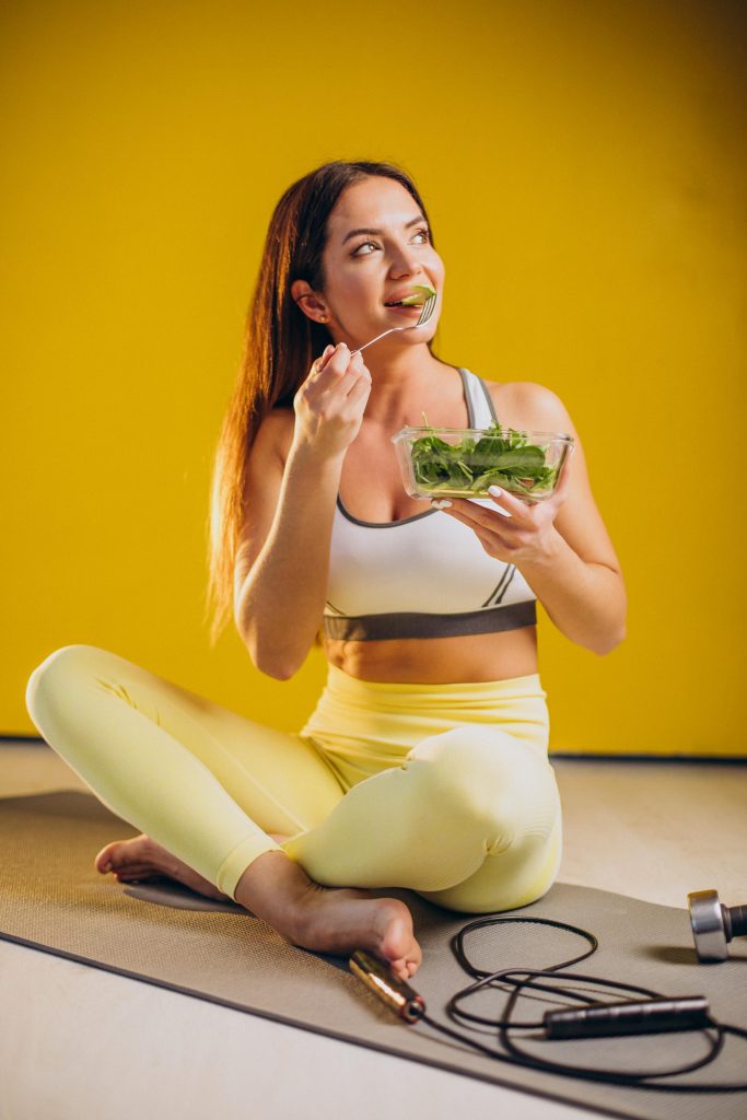 Imagem de uma mulher sentada em cima de um tapete  de ioga com as pernas cruzadas e roupa de malhação. Ela segura uma  vasilha com salada e leva um garfo à boca