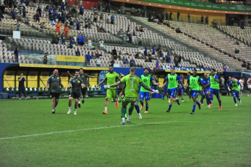Imagem do goleiro do Nacional comemorando de costas pra câmera, enquanto jogadores do time correm na direção dele. O goleiro usa um uniforme verde limão e os jogadores de linha colete verde limão e calção azul