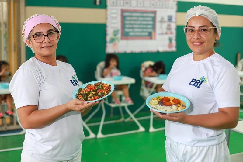 Imagem de duas merendeiras vestidas com camisa e calça branca. Elas usam uma touca de pano na cabeça e seguram pratos de comida. Ao fundo, em desfoque, crianças estão sentadas em cadeirinhas de alimentação