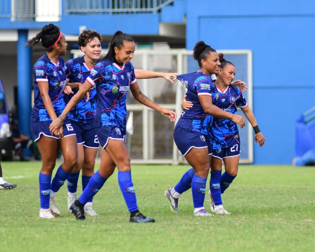 Jogadoras do 3B, com uniforme azul, com detalhes em branco e rosa abraçam jogadora, comemorando gol