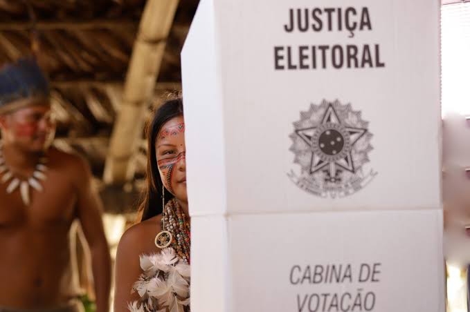 Mulher indígena atrás de uma cabine de votação da Justiça Eleitoral durante votação.